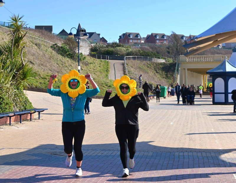 Image shows two runners in daffodil hats smiling with fists in the air at Barry County Councils annual fun run. The town of Barry can be seen in the background with a bright blue sky and fields of green.