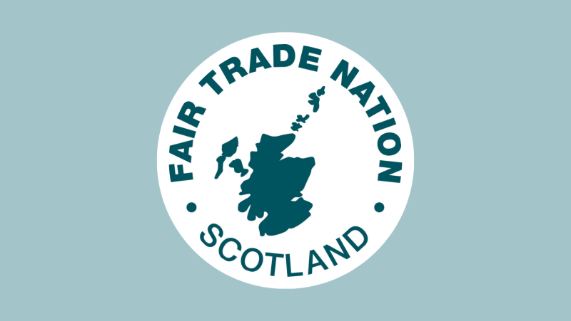 Photo depicts the 'Fair Trade Nation Scotland' Emblem with the Scotland map outline in the middle of the picture in dark green all on a white circle. This circle is on a plain pale blue background.