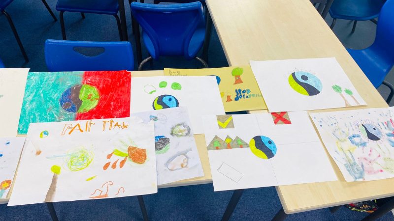Image shows lots of drawings created by students of what they think about Fair Trade. Pictures are on a brown classroom table with a blue carpet and blue school chairs in the background. Many children have used the Fair Trade logo in their artwork, which is a blue, green, black and white in a circle.